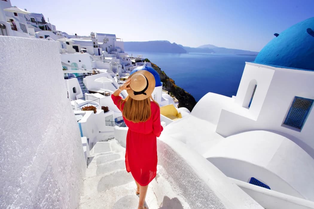 Beautiful girl in red dress and hat comes down the stairs exploring Oia village - Santorini